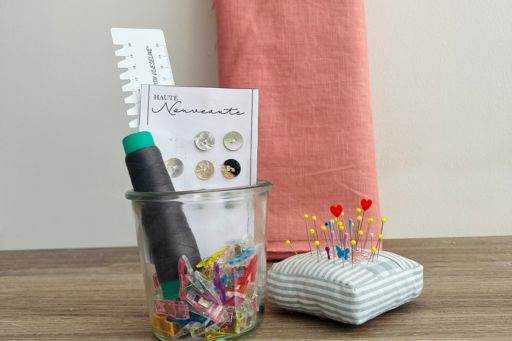 Arrangement von Nähzubehör auf einem Holztisch. Es sind eine graue Stoffrolle, eine kleine Glasschale mit bunten Klammern, eine Packung Knöpfe, ein Lineal, eine Nadelkissen in einem gestreiften Stoff mit bunten Stecknadeln, eine Schere und ein Stück korallenfarbener Stoff zu sehen. Die Gegenstände sind für eine Näharbeit vorbereitet und der Hintergrund ist eine helle Wand